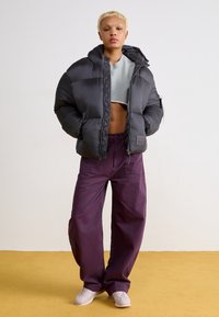 Giacca a vento nera con cappuccio, caratterizzata da una vestibilità corta e texture trapuntata. Abbinata a pantaloni viola larghi e sneakers chiare.