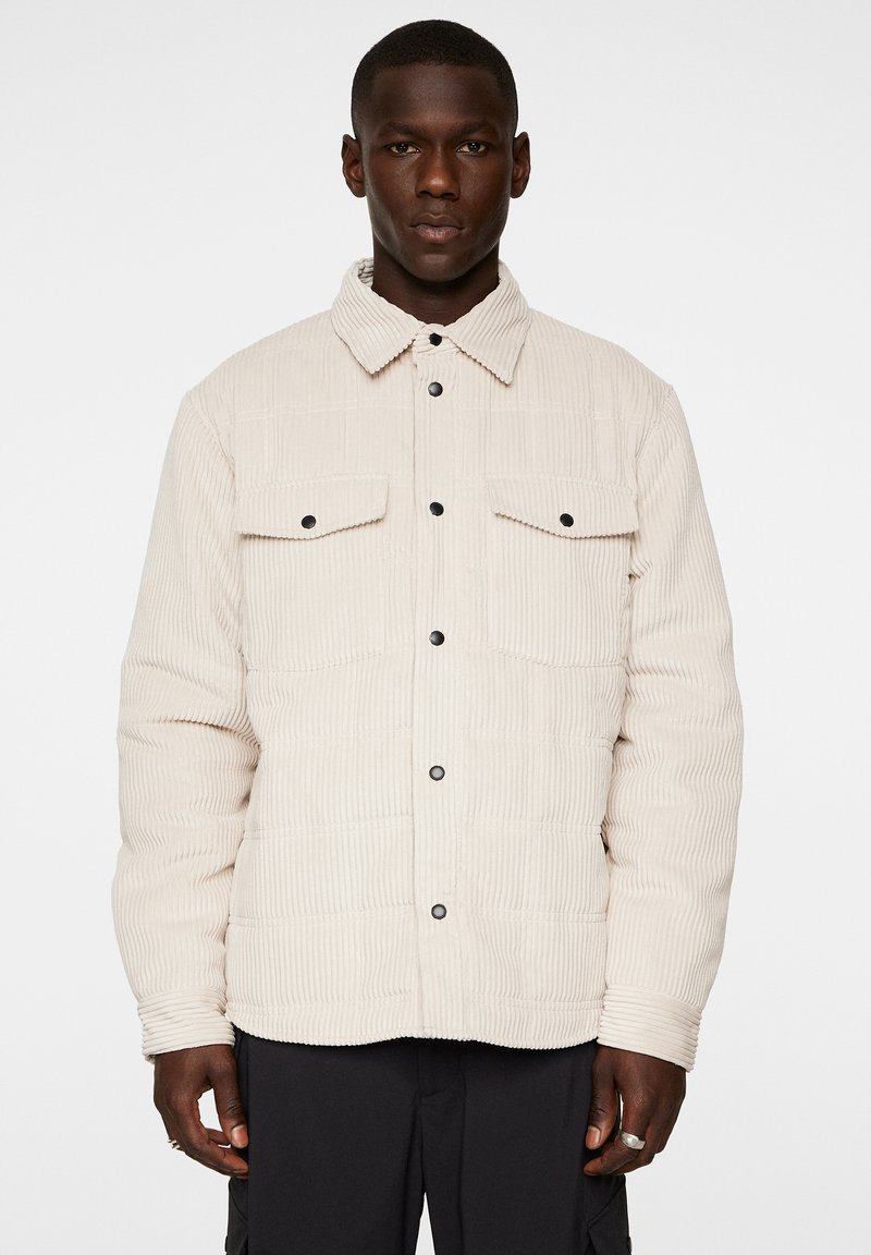 Beige kordjacka med knappfäste framtill, två bröstfickor och krage; har svarta knappar och textil med struktur.