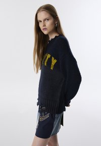 Pull en maille bleu marine avec un ourlet effiloché et "UNITY" en lettres jaune vif. Assorti à une jupe en denim avec des accents dorés.