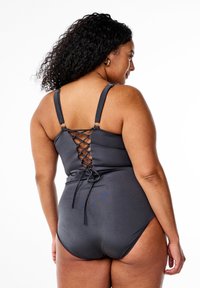 Viens gabals peldkostīms tumši pelēkā krāsā ar izsieta aizmuguri, regulējamām siksnām un mīkstu tekstūru. Iekļauj dekoratīvu loku detaļu.