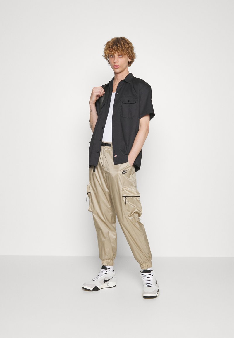 Chemise noire à manches courtes, pantalon cargo beige avec zippers, et baskets blanches avec accents noirs. Texture lisse, design utilitaire.