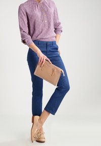 Woman wearing a purple patterned blouse and blue pants holds a beige clutch, standing with crossed legs in beige tassel loafers.