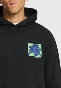 Felpa nera con finitura texturizzata. Presenta un patch grafico verde e blu che rappresenta una mano stilizzata. Tasca a canguro sul davanti.
