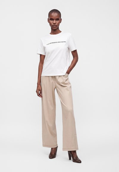 Femme portant un t-shirt blanc avec un texte, un pantalon beige à jambes larges et des chaussures marron à talons, debout avec une main dans la poche sur fond blanc.