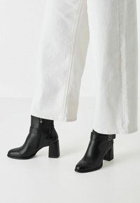 Bottines en cuir noir à bout pointu et talon carré, dotées d'une fermeture éclair sur le côté et d'une sangle avec un accent métallique circulaire.