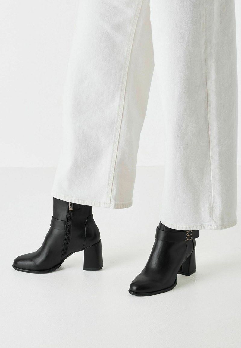 Bottines en cuir noir à bout pointu et talon carré, dotées d'une fermeture éclair sur le côté et d'une sangle avec un accent métallique circulaire.