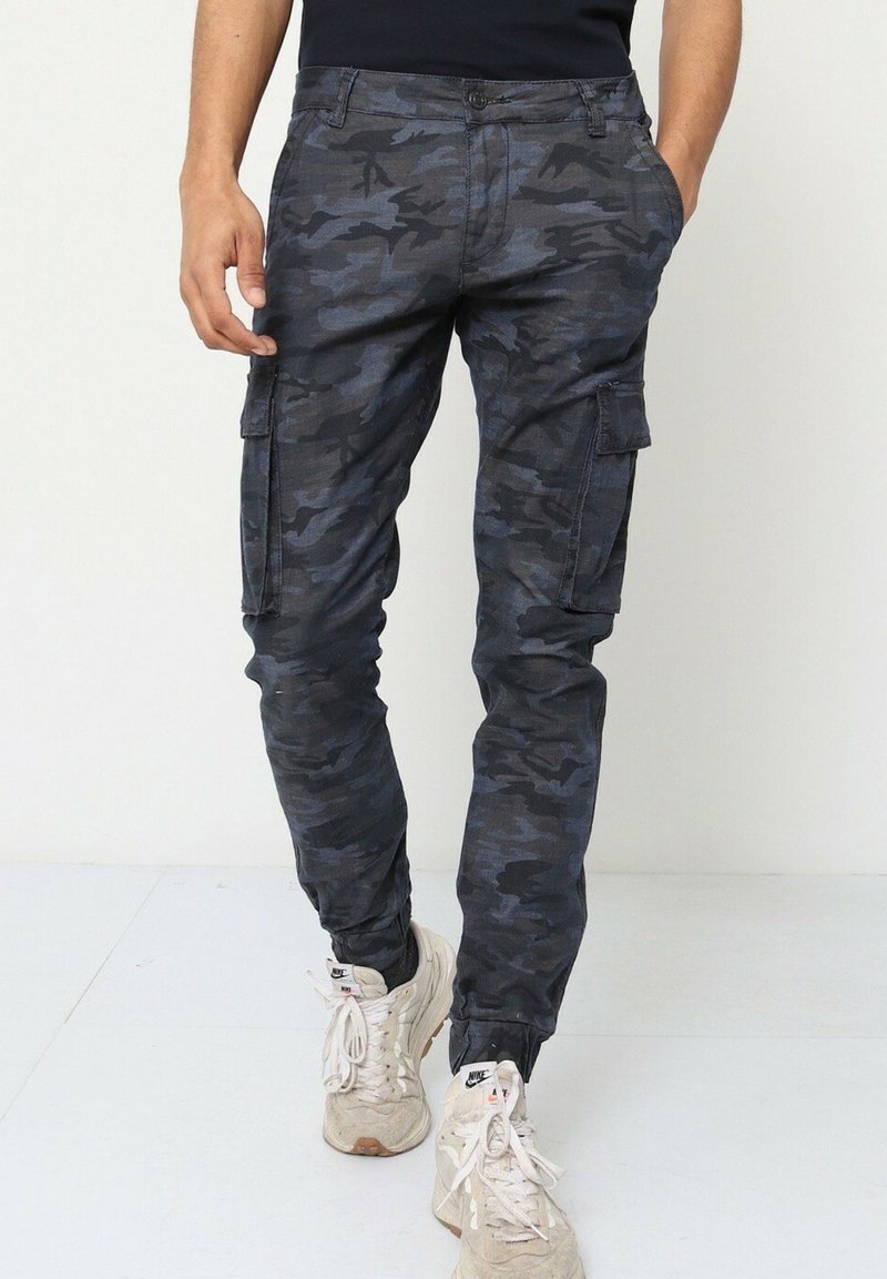 Pantalons cargo camouflés en tissu noir et gris foncé, dotés de poches latérales, de coupe slim et d'une texture lisse. Associés à des baskets neutres.