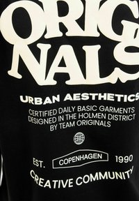Černé bavlněné tričko s velkým bílým textem: "ORIGINALS URBAN AESTHETICS" a dalšími brandingovými detaily níže, včetně ikony zeměkoule.