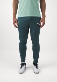 Nike vihreät urheiluhousut, joissa on kiinteä malli, sileä pinta ja logoyksityiskohta reidessä, yhdistettynä valkoisiin tennareihin.
