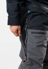 Hand unzipping side ventilation on gray and black Swedemount outdoor pants with Swedish flag tab visible.