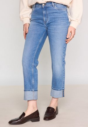 Hellblaue, hochgeschnittene Jeans mit geradem Schnitt, die umgeschlagene Säume und eine ausgewaschene Textur aufweisen. Kombiniert mit dunkelbraunen Loafers.
