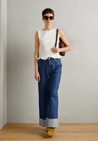 Vit texturerad ärmlös topp, blå hög midja vida jeans med ljus denimuppvik, och bruna klackstövlar, accessoriserat med solglasögon.