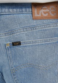 Ljust blå denimjeans har ett texturerat tyg, synliga sömmar och en brun läderlapp med präglad "Lee"-logotyp vid midjan.