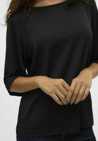 Top negro texturizado con un corte holgado, escote redondeado y mangas de tres cuartos. Manos colocadas cerca de la cintura, uñas esmaltadas en un color neutro.