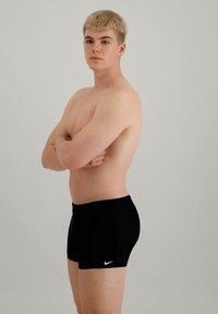 Svarte badeshorts med Nike-logo, tettsittende design, flat midjebånd og jevn tekstur, båret av en person med armene krysset i en nøytral positur.