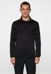 Svart zip-up jacka med hög krage och långa ärmar. Tillverkad av slät tyg, med en diskret logotyp på bröstet och ärmsluten.