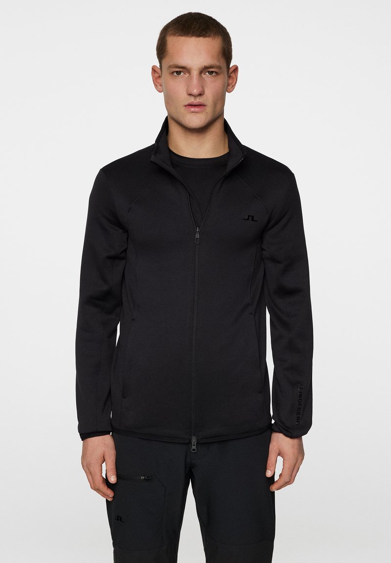 Svart zip-up jacka med hög krage och långa ärmar. Tillverkad av slät tyg, med en diskret logotyp på bröstet och ärmsluten.