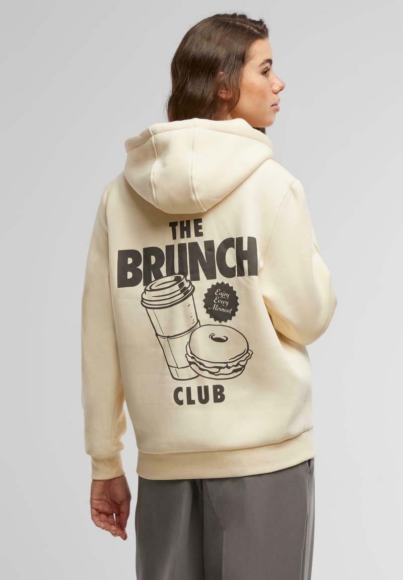 Krémová mikina s kapucňou a potlačou na chrbte, ktorá zobrazuje šálku kávy, hamburger a text "THE BRUNCH CLUB." Mäkká, pohodlná tkanina, predná kengurú kapsa.
