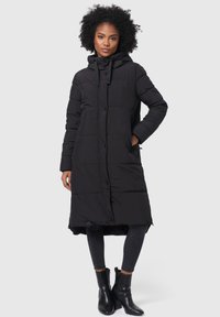Abrigo de puff negro con capucha, de longitud larga, diseño acolchado, botones delanteros y bolsillos laterales, combinado con jeans oscuros y botas de tobillo.