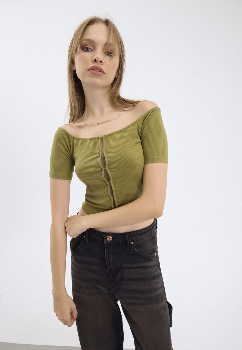 Olivovo zelený rebrovaný crop top s dizajnom bez ramienok a detailom predného gombíka, kombinovaný s čiernymi džínsami s vysokým pásom a jemným vyblednutím.