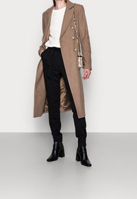 Bruine wollenblend jas met double-breasted ontwerp, zwarte broek en zwarte enkellaarsjes met hak. Gelaagde witte gebreide trui eronder.