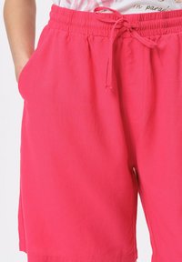Różowe bawełniane szorty z elastycznym paskiem w talii, regulowanym sznurkiem i bocznymi kieszeniami. Gładka tekstura i długość do kolan.