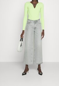 Rippenshirt in Neon Grün mit langen Ärmeln, V-Ausschnitt und Schnürdetails, kombiniert mit hellgrauen, weiten Jeans und schwarzen spitzen High Heels.