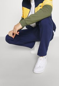 Personne portant une veste zippée jaune, bleu marine et olive, un pantalon bleu marine et des baskets blanches, accroupie avec les mains jointes de façon détendue.