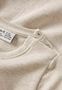 Helles beiges Baumwollhemd mit Rundhalsausschnitt. Verfügt über Druckknopfverschlüsse und gerippte Bündchen. Pflegeetikett auf dem Stoff sichtbar. Weiche Textur.