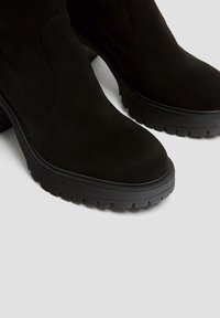 Botas de tobillo de ante negras con una suela de goma gruesa, punta redondeada y paneles elásticos laterales para facilitar su uso. Superficie texturizada con un profundo dibujo.
