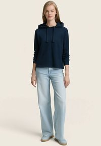 Námořnická mikina s kapucí v uvolněném střihu, s žebrovanými manžetami a stahovací šňůrkou. Doplněná světle modrými džíny se střihnutými nohavicemi a béžovou obuví.