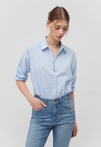 Hellblaues Hemd mit kleinen gemusterten Punkten, hochgerollten Ärmeln und klassischem Kragen, kombiniert mit hoch sitzenden Jeans.