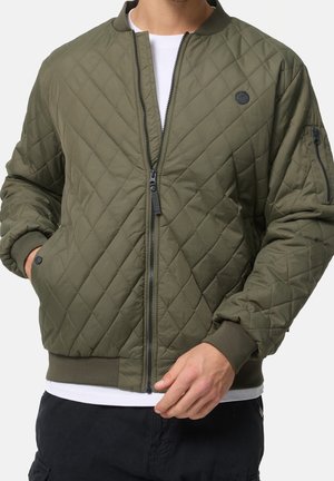 Mężczyzna w zielonej pikowanej kurtce bomber z zamkiem z przodu, ściągaczami na rękawach i małym logo na piersi, na białej koszuli.