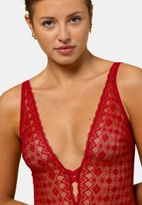 Body en dentelle rouge avec un décolleté en V profond, des motifs floraux et des bretelles ajustables avec des accents croisées à l'avant.