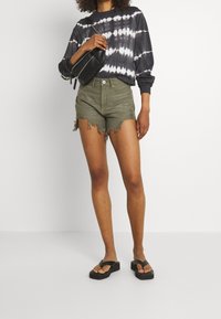 Camisa de manga larga negra con efecto de teñido anudado, pantalones cortos deshilachados en verde oliva, bolso de cuero negro tipo bandolera y sandalias de plataforma negras. Outfit casual.