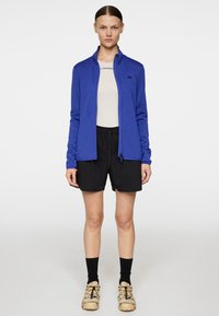 Giacca blu con zip e maniche lunghe, abbinata a una maglia beige, pantaloni corti neri, calze nere e calzature beige con accenti colorati.