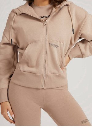 Frau trägt beigen Kapuzenpullover mit Reißverschluss und GUESS-Logo sowie passende beige Leggings mit teilweise sichtbarem Logo am Oberschenkel.