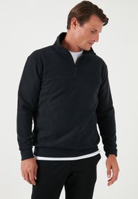 Pullover in pile nero con colletto a zip a quarto, polsini e orlo a costine. Texture morbida e finitura liscia, abbinato a una maglietta bianca.