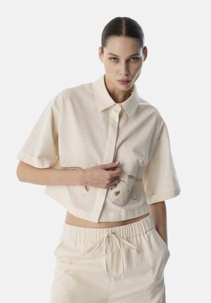 Kurzes, cremefarbenes, cropped Shirt mit einem Button-Down-Kragen und kurzen Ärmeln, das über feine Cut-Out-Stickereien am Saum verfügt.
