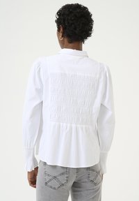Valkoinen pitkähiha blouse, jossa on rypytetty selkä, pleattyjä yksityiskohtia ja kevyesti levenevät hihansuut. Yhdistetty harmaisiin farkkuihin, joissa on koristeellista tikkausta.