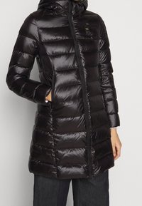 Czarna, błyszcząca pikowana długa kurtka puchowa z przednim zamkiem, wysokim kołnierzem, kieszenią na łatkę i rękami w bocznych kieszeniach, noszona z ciemnymi spodniami.