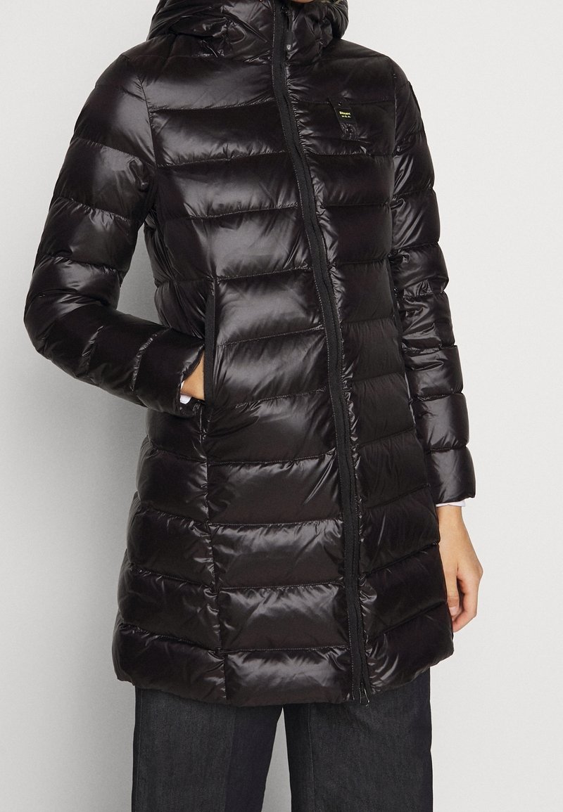 Czarna, błyszcząca pikowana długa kurtka puchowa z przednim zamkiem, wysokim kołnierzem, kieszenią na łatkę i rękami w bocznych kieszeniach, noszona z ciemnymi spodniami.