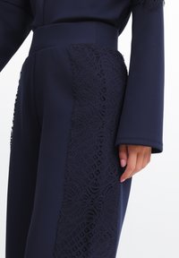 Pantaloni bleumarin cu talie ajustată și panou lateral cu detalii complexe din dantelă. Țesătura este netedă, cu un finisaj mat.