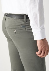 Slim-fit, olive green trousers with a smooth texture, featuring a back pocket with a button and a leather belt. Label visible on the pocket.