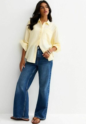 Kobieta ubrana w luźną, bladożółtą koszulę na guziki, niebieskie szerokie spodnie dżinsowe, brązowe sandały i bransoletki, stojąca przy białej ścianie.