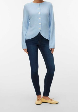 Femme portant un cardigan bleu clair boutonné, un jean skinny foncé et des ballerines beiges, debout devant un fond blanc.
