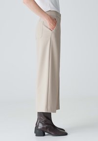 Pantalons beiges taille haute avec une coupe ample et une poche arrière unique, associés à des bottes à talons marron foncé et un simple haut blanc.