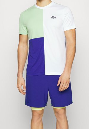 Mand iført en farveblokeret t-shirt i grøn, hvid og blå med blå shorts og gule underbukser, står med afslappede arme.