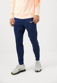 Navy athletic pants with a fitted design, featuring a pink Nike logo and a Barcelona crest. Worn with light gray Nike shoes.