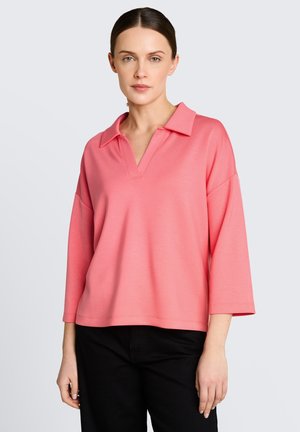 Kobieta ubrana w luźną różową bluzkę na długi rękaw z szerokim kołnierzem i czarne spodnie, stojąca na jednolitym jasnym tle.